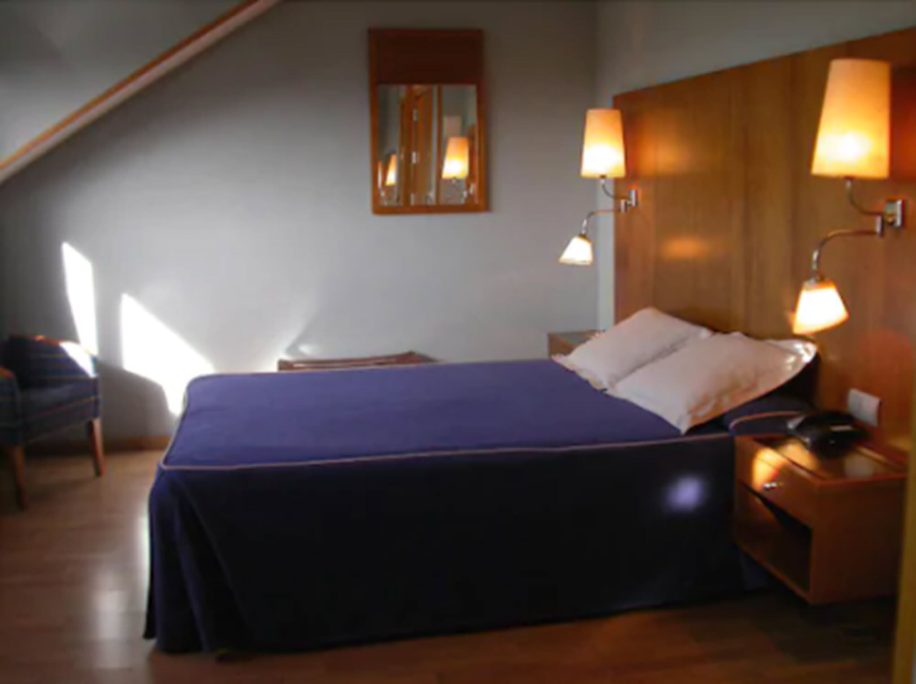 Un dormitorio con cama, lámparas de noche, mesita de noche, una silla y una ventana.