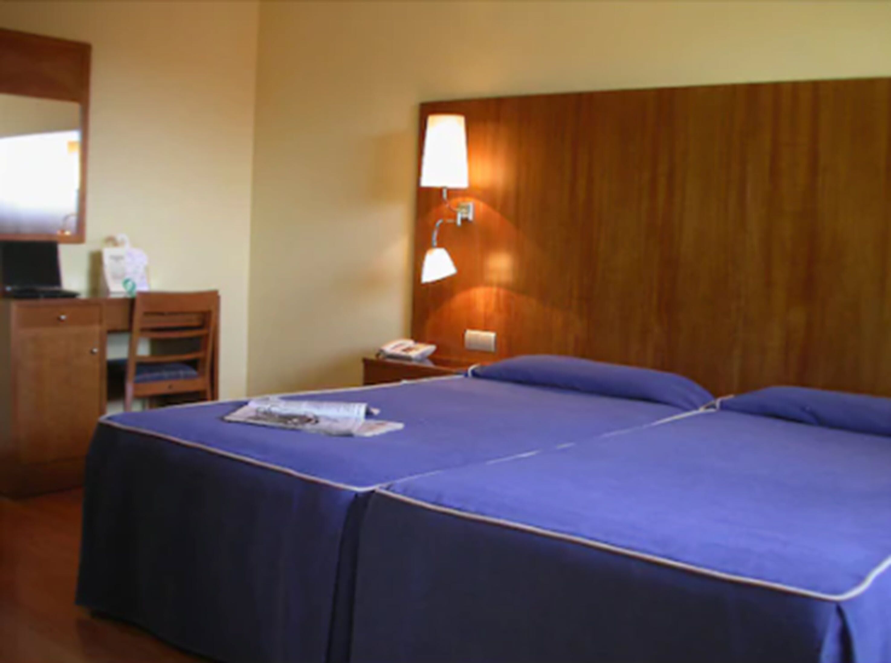 A hotel room with a wooden headboard, a desk with a lamp, a chair, and a bed with a blue bedspread.