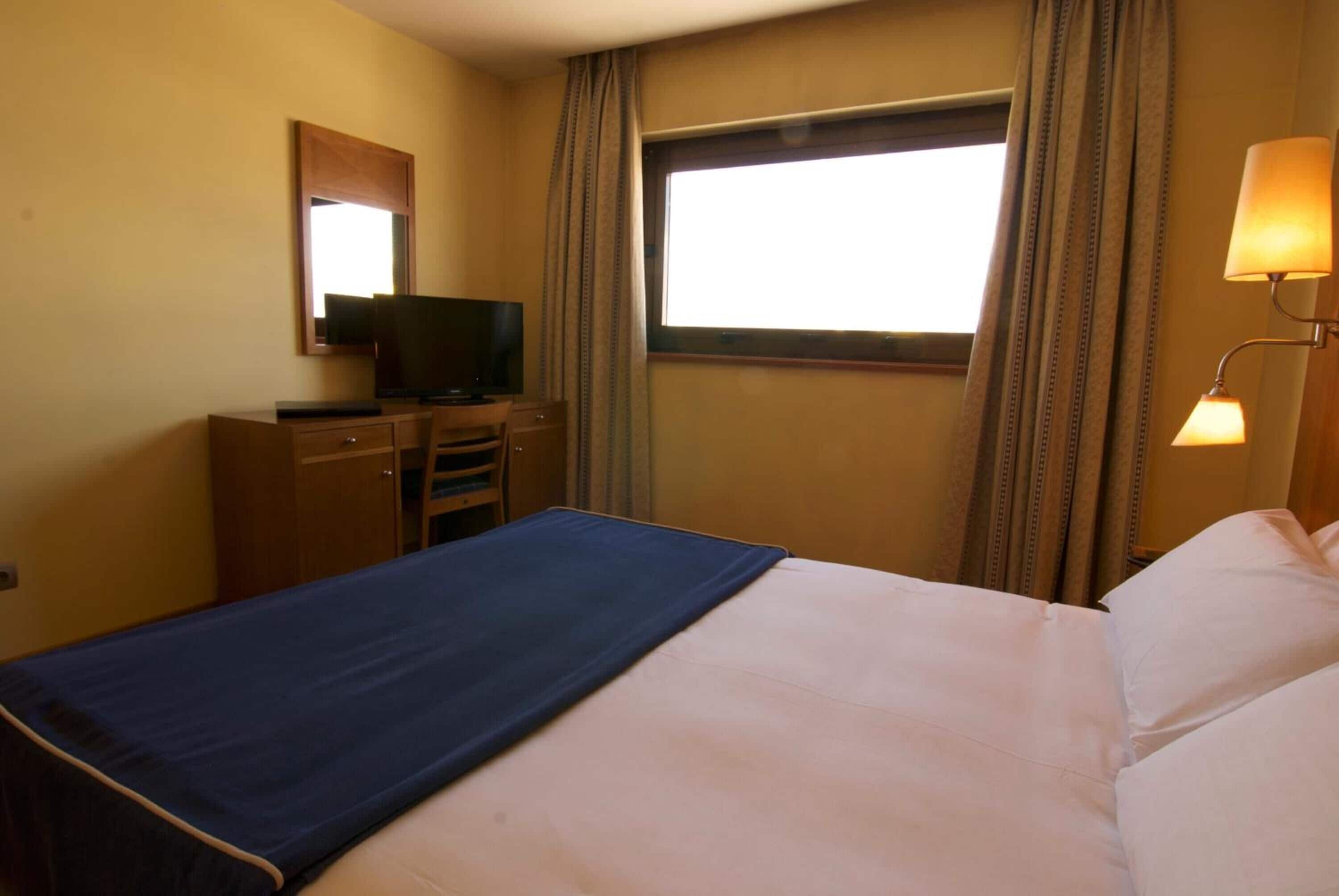 Habitación de hotel con cama, escritorio con televisor, ventana con cortinas y lámpara.