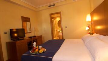 A hotel room with a bed, a television, a desk with a tray of fruit, and a nightstand with a lamp.
