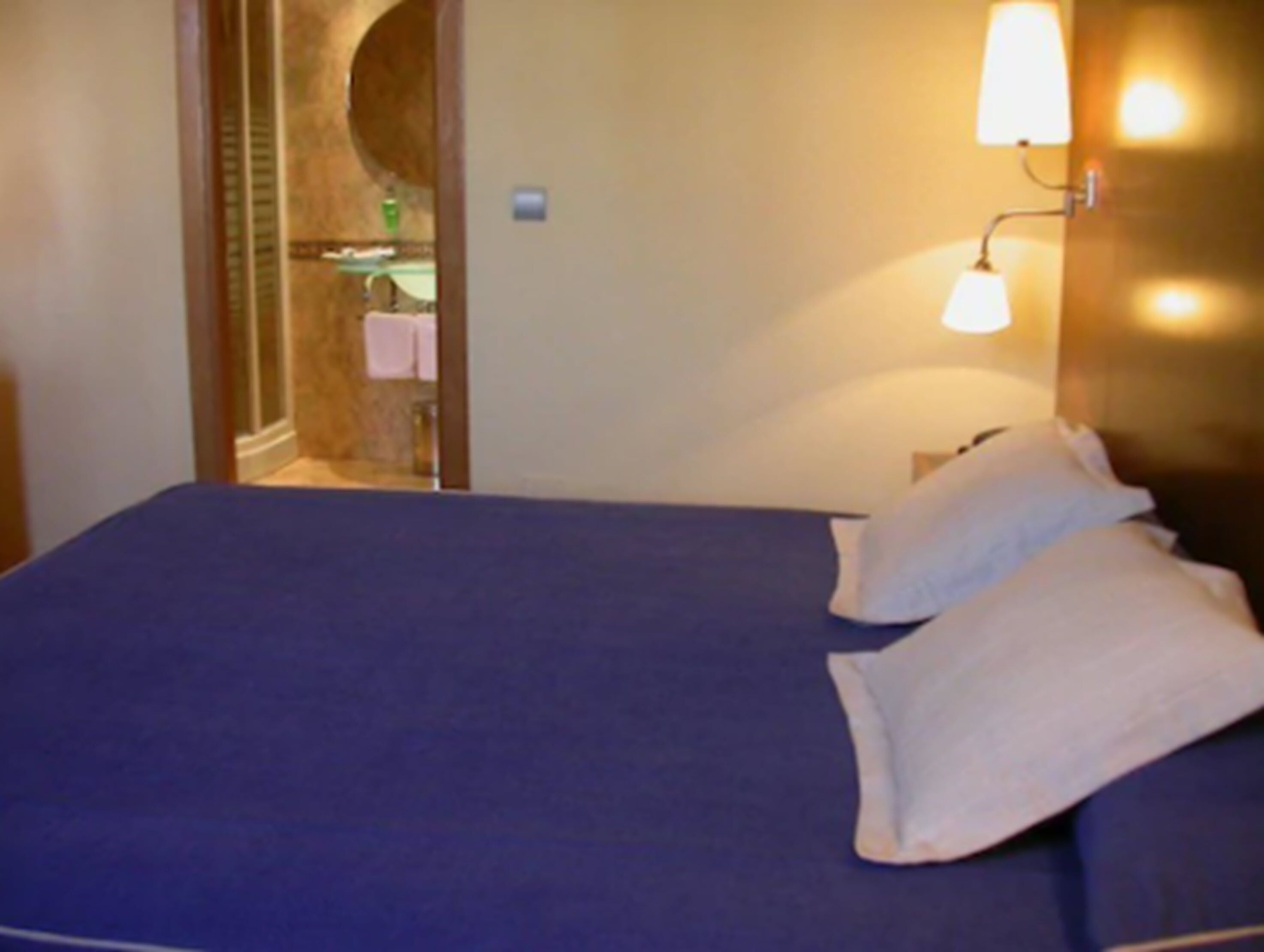 A hotel room with a bed, a nightstand, a wall-mounted lamp, and a bathroom visible through an archway.