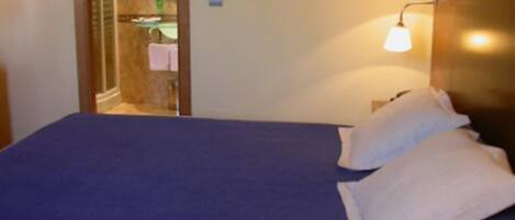 A hotel room with a bed, a nightstand, a wall-mounted lamp, and a bathroom visible through an archway.