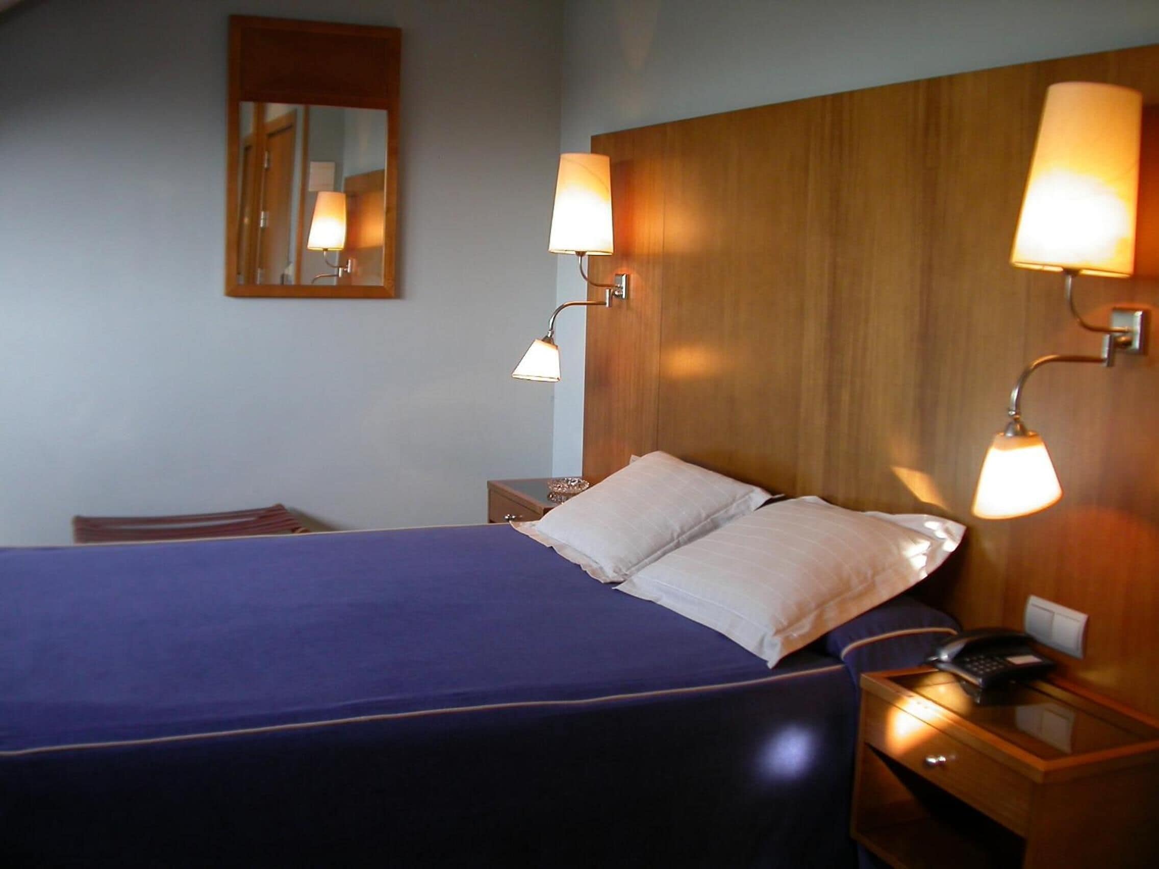Una habitación de hotel con una cama, dos lámparas de noche, una mesita de noche con un teléfono y un espejo en la pared.