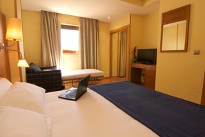 Une chambre d’hôtel avec un lit, un bureau avec un ordinateur portable, une télévision, une chaise et une fenêtre avec des rideaux.