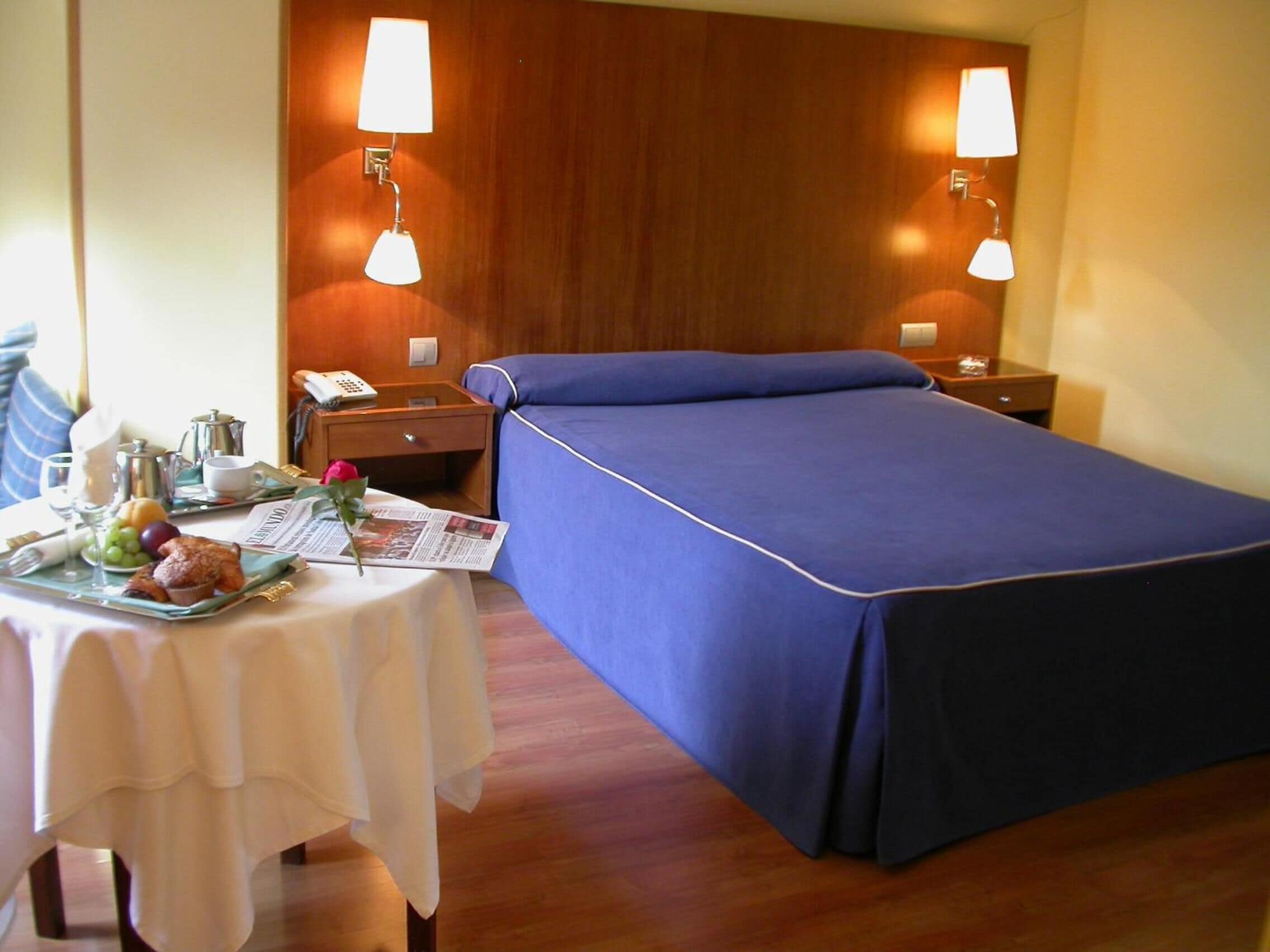 Quarto de hotel com uma cama bem arrumada, mesa de cabeceira com abajur, pequena mesa com itens de café da manhã e cabeceira de madeira.