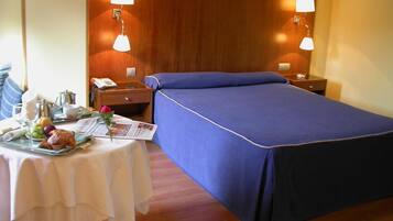 Quarto de hotel com uma cama bem arrumada, mesa de cabeceira com abajur, pequena mesa com itens de café da manhã e cabeceira de madeira.