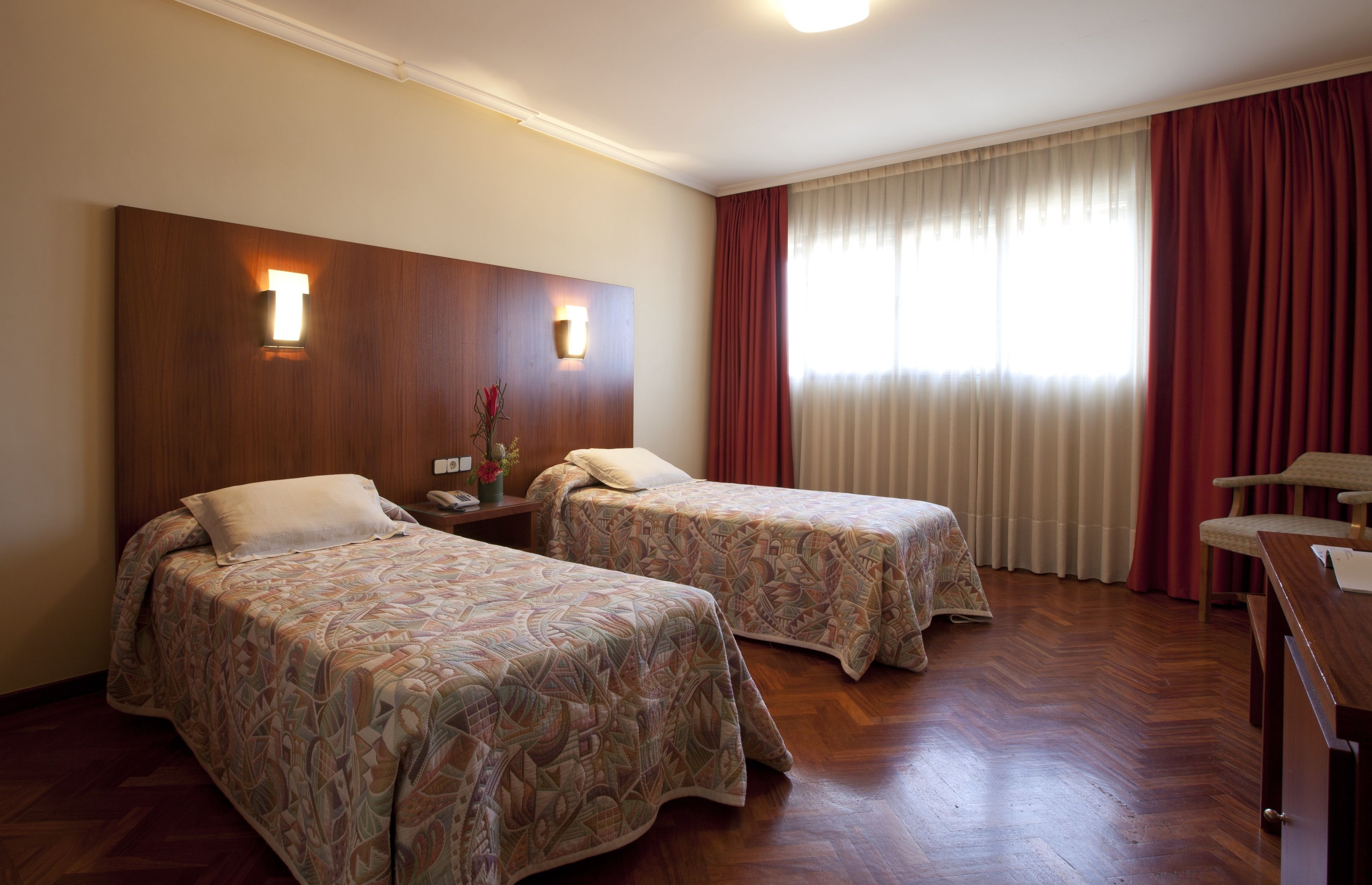 Habitación de hotel con dos camas, cabecera de madera, escritorio y ventana con cortinas.
