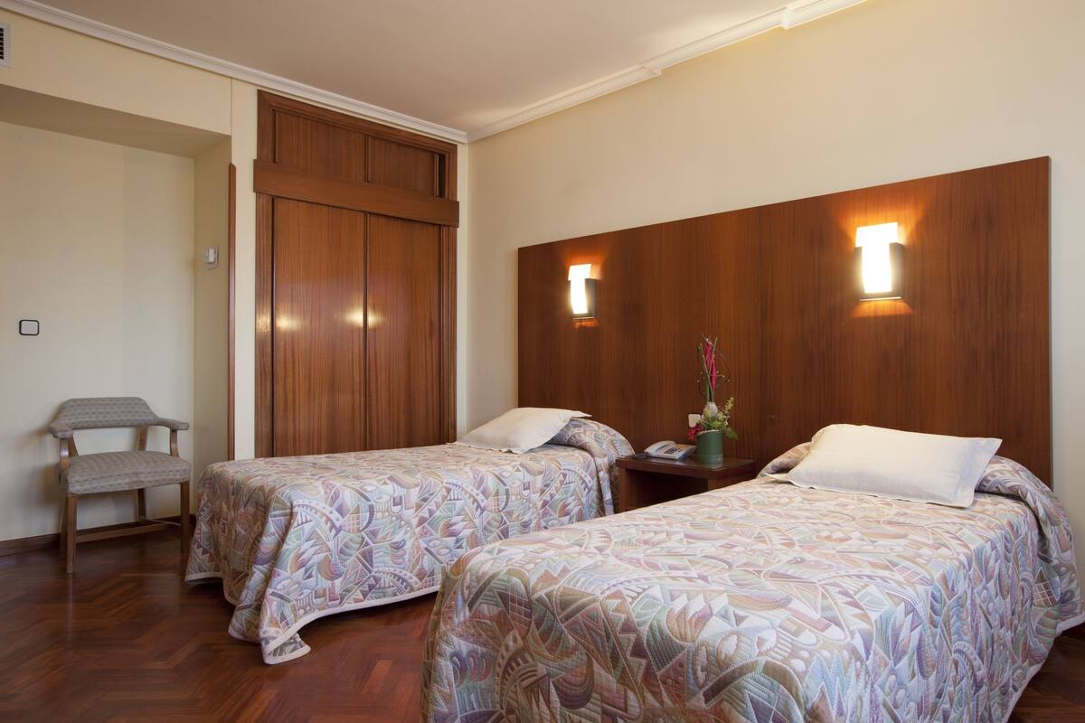 Habitación de hotel con dos camas individuales, cabecera de madera, mesita de noche con teléfono y florero, y una silla con cojín.