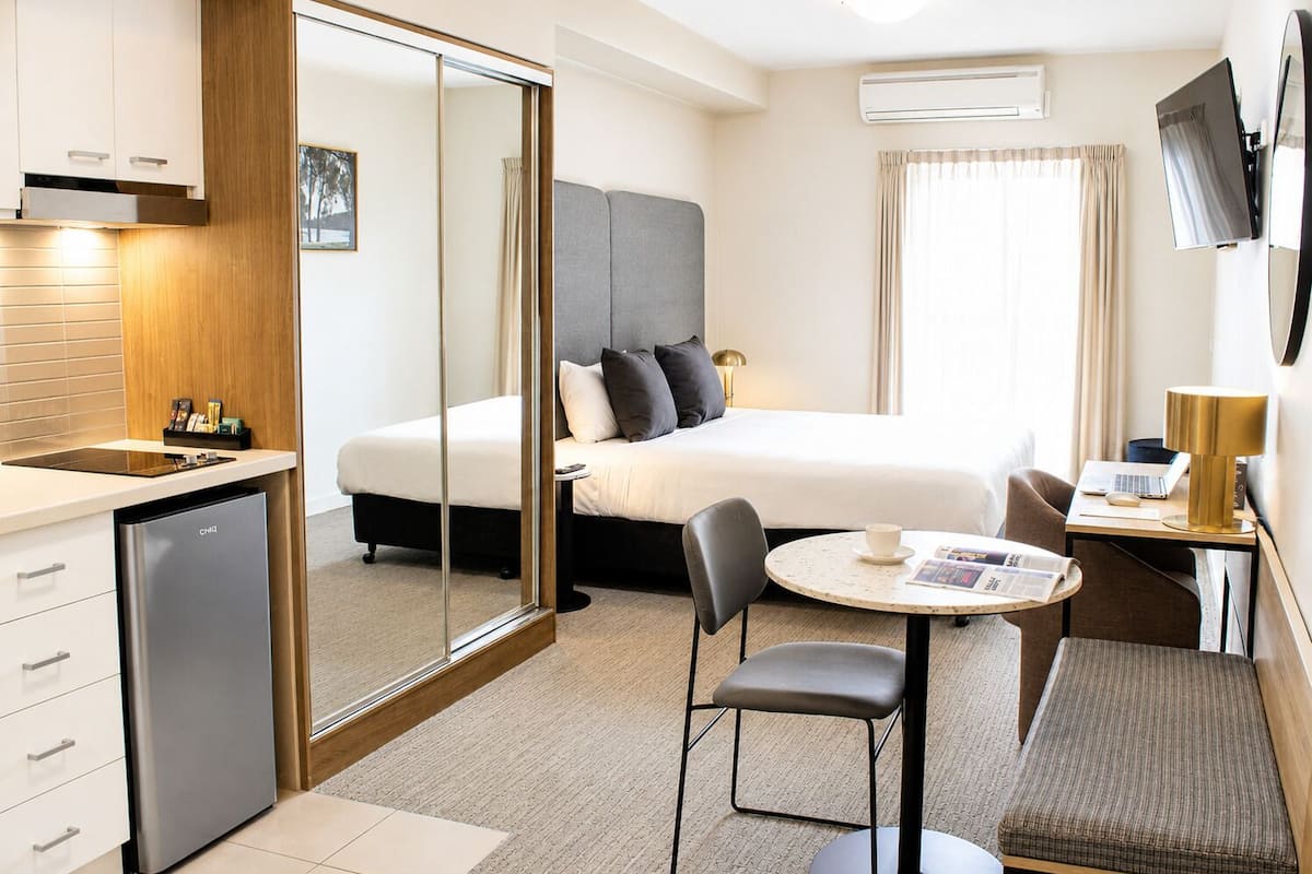Ein modernes Hotelzimmer mit einem Bett, einem Schreibtisch mit einem Laptop, einem Sessel, einem kleinen Tisch mit einer Tasse, einem Kühlschrank und einem an der Wand montierten Fernseher.