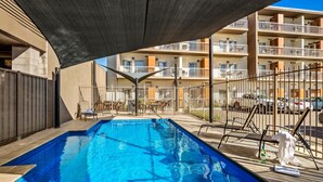 A swimming pool with a shaded area, surrounded by a fence, and adjacent to a multi-story building with balconies. - Quest Mildura (Mildura)