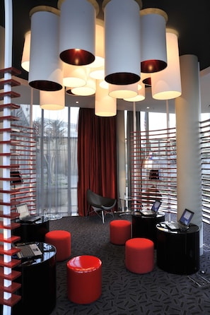 A modern lounge area with a red and black color scheme, featuring a seating area with ottomans, a laptop on a table, and a large window with blinds. - Park Inn Sandton (Sandton)