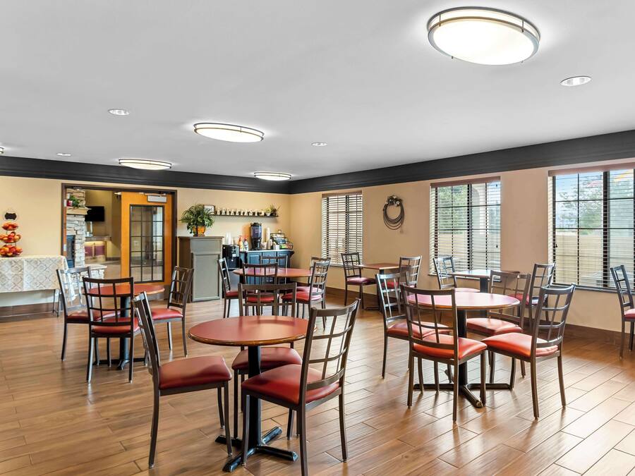 A spacious dining area with round tables and chairs, a kitchen in the background, and large windows with blinds.