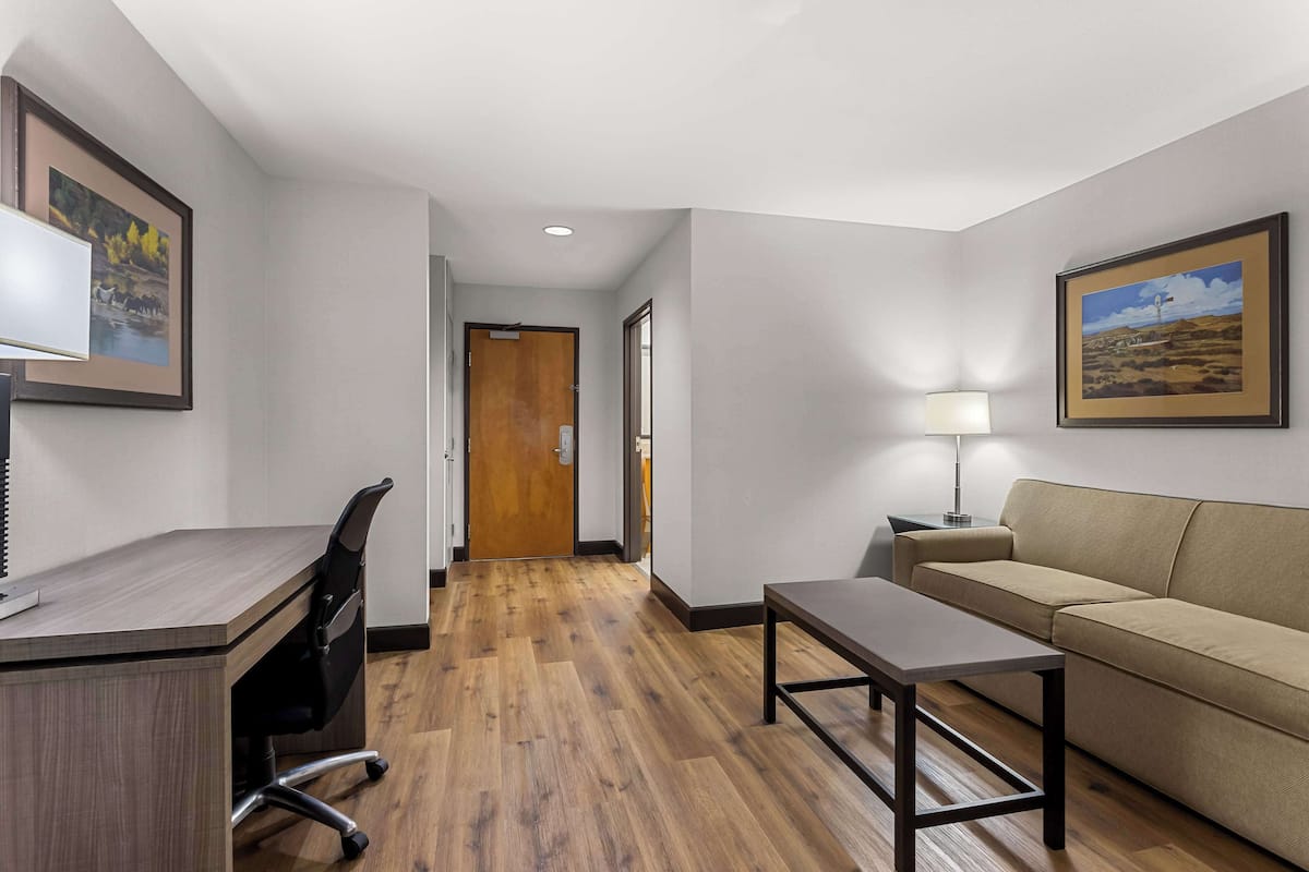 A hotel room with a wooden desk, a black office chair, a beige sofa, a small table, and a lamp. There is a door leading to another room and a framed picture on the wall.