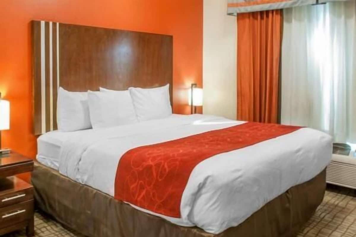 A hotel room with a large bed, a wooden headboard, a red accent on the bedspread, a nightstand with drawers, and a window with curtains.