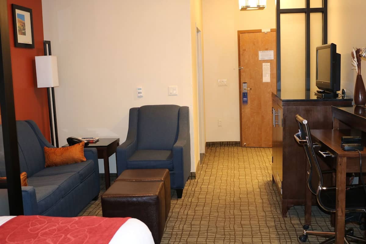 A hotel room with a bed, a blue armchair, a small table with a lamp, a wooden desk with a computer, and a door.