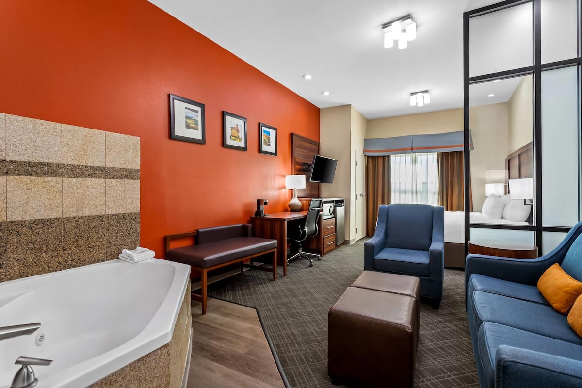 A hotel room with a large bed, a sitting area with a sofa and armchair, a desk with a chair, a television, and a bathroom with a bathtub and sink.
