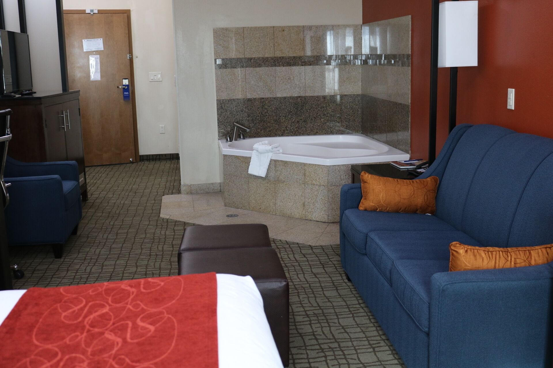 A hotel room with a large bed, a blue sofa, a brown ottoman, a small table, a desk with a computer, a television, a wooden door, a bathroom with a bathtub, and a mirror.