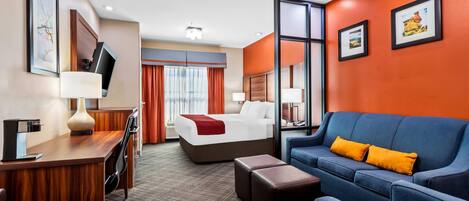 Une chambre d’hôtel avec un lit, un bureau et une chaise, un canapé bleu avec des coussins orange, et une grande fenêtre avec des rideaux.