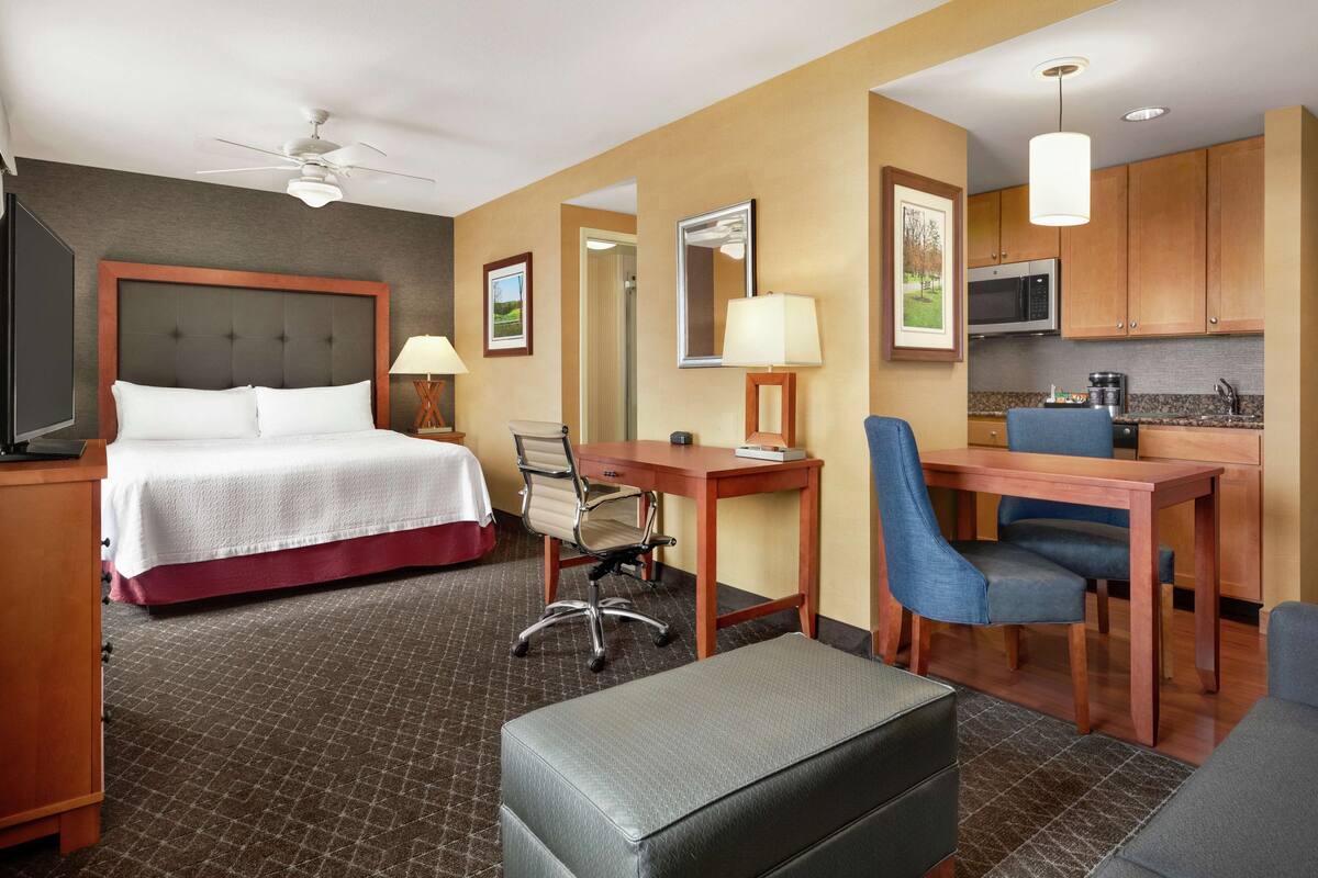 Ein Hotelzimmer mit Bett, Schreibtisch, Stuhl, Küchenzeile und Deckenventilator.