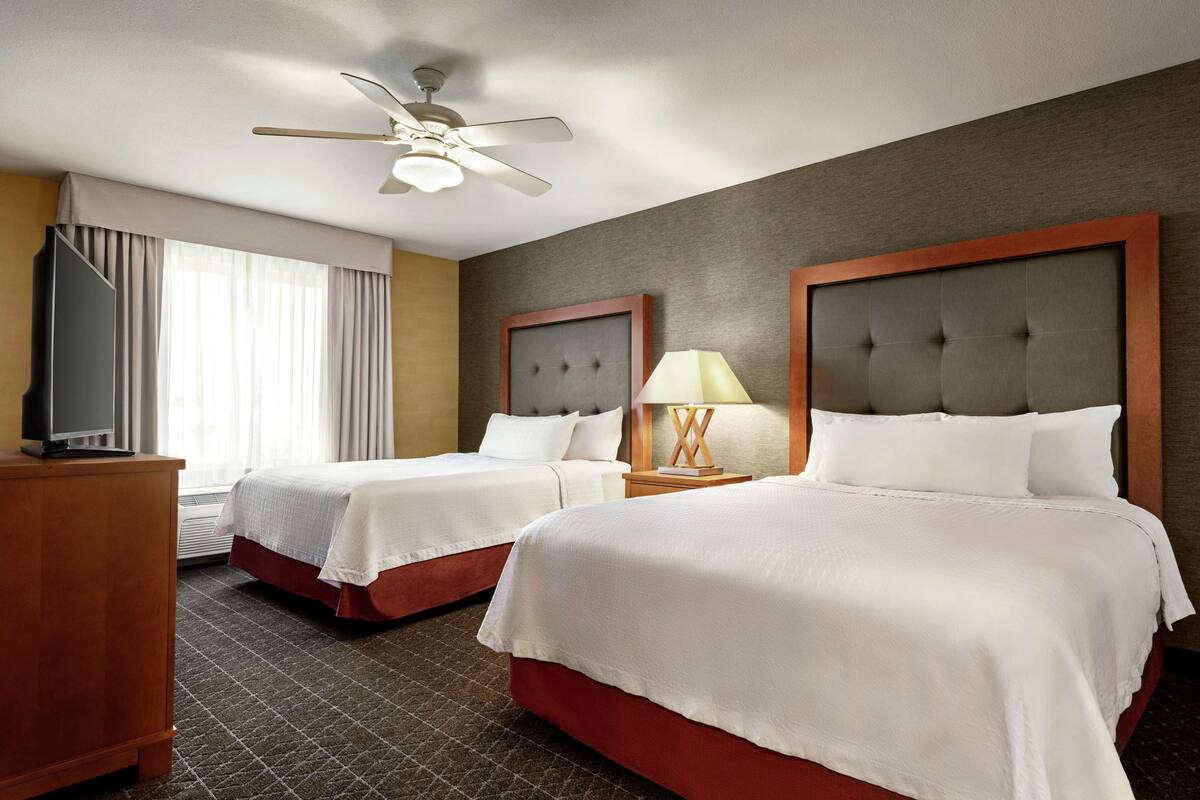 Ein Hotelzimmer mit zwei Betten, einem Deckenventilator, einem Fernseher auf einem Holzkasten und einem Fenster mit Vorhängen.