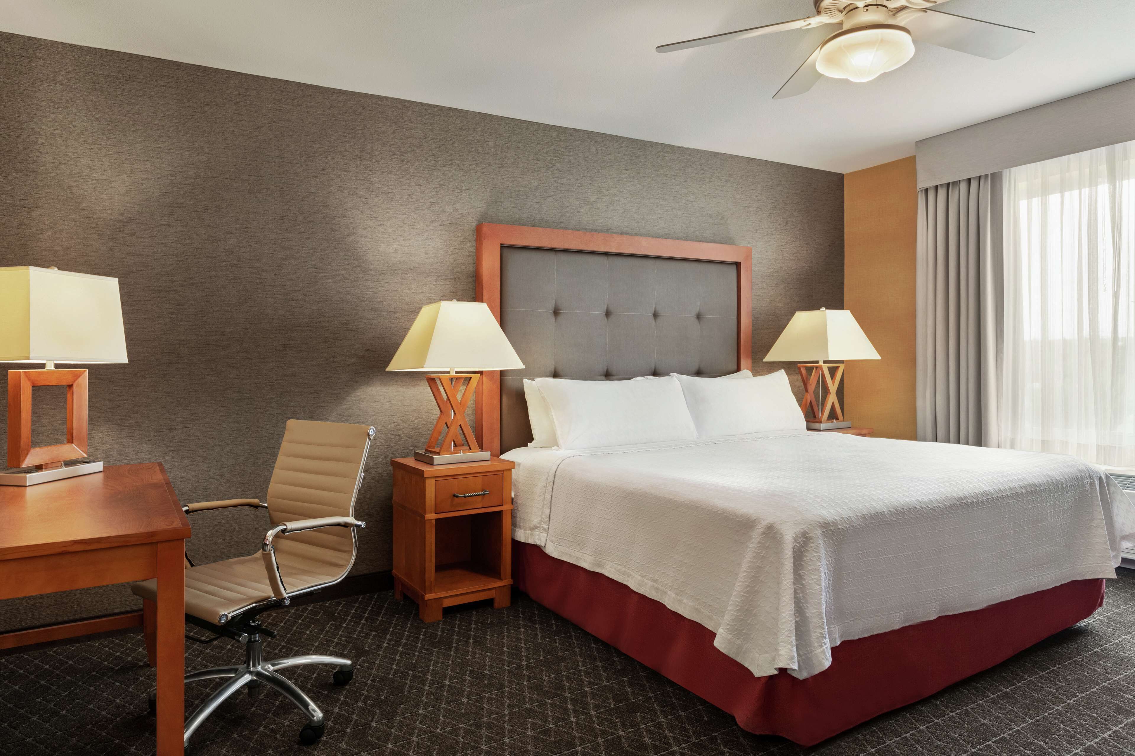 Una habitación de hotel con una cama grande, dos mesitas de noche con lámparas, una mesita de madera, una silla y un ventilador de techo.