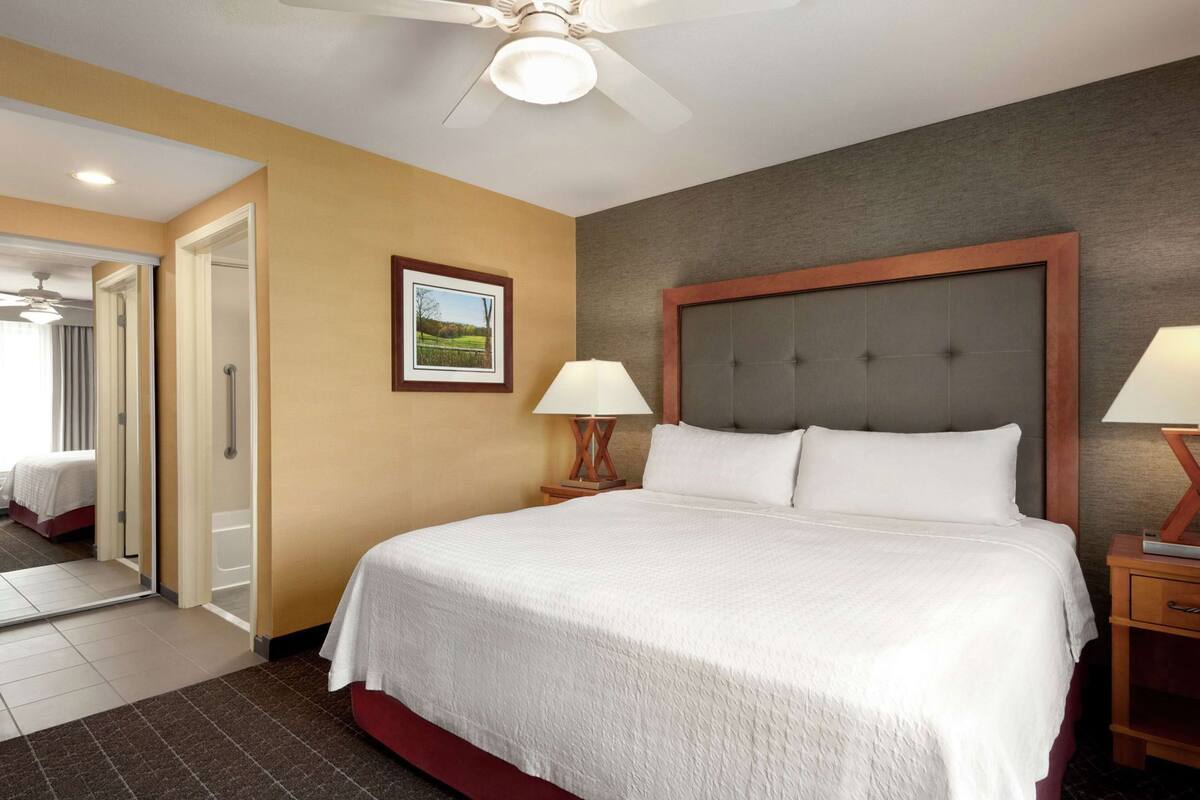 Ein Hotelzimmer mit einem großen Bett, einem Deckenventilator, einem gerahmten Bild an der Wand und einem Nachttisch mit einer Lampe.