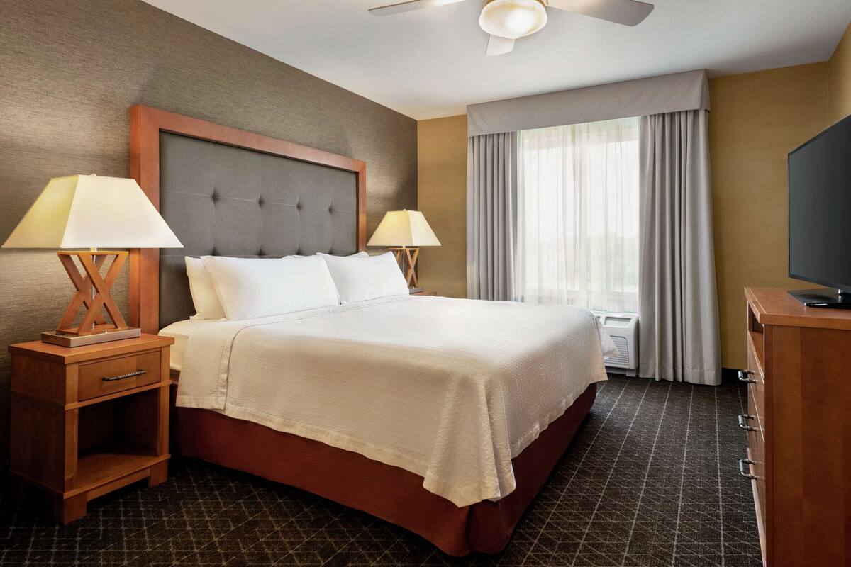 Ein Hotelzimmer mit einem großen Bett, zwei Nachttischen inklusive Lampen, einem Flachbildfernseher und einem Fenster mit Vorhängen.