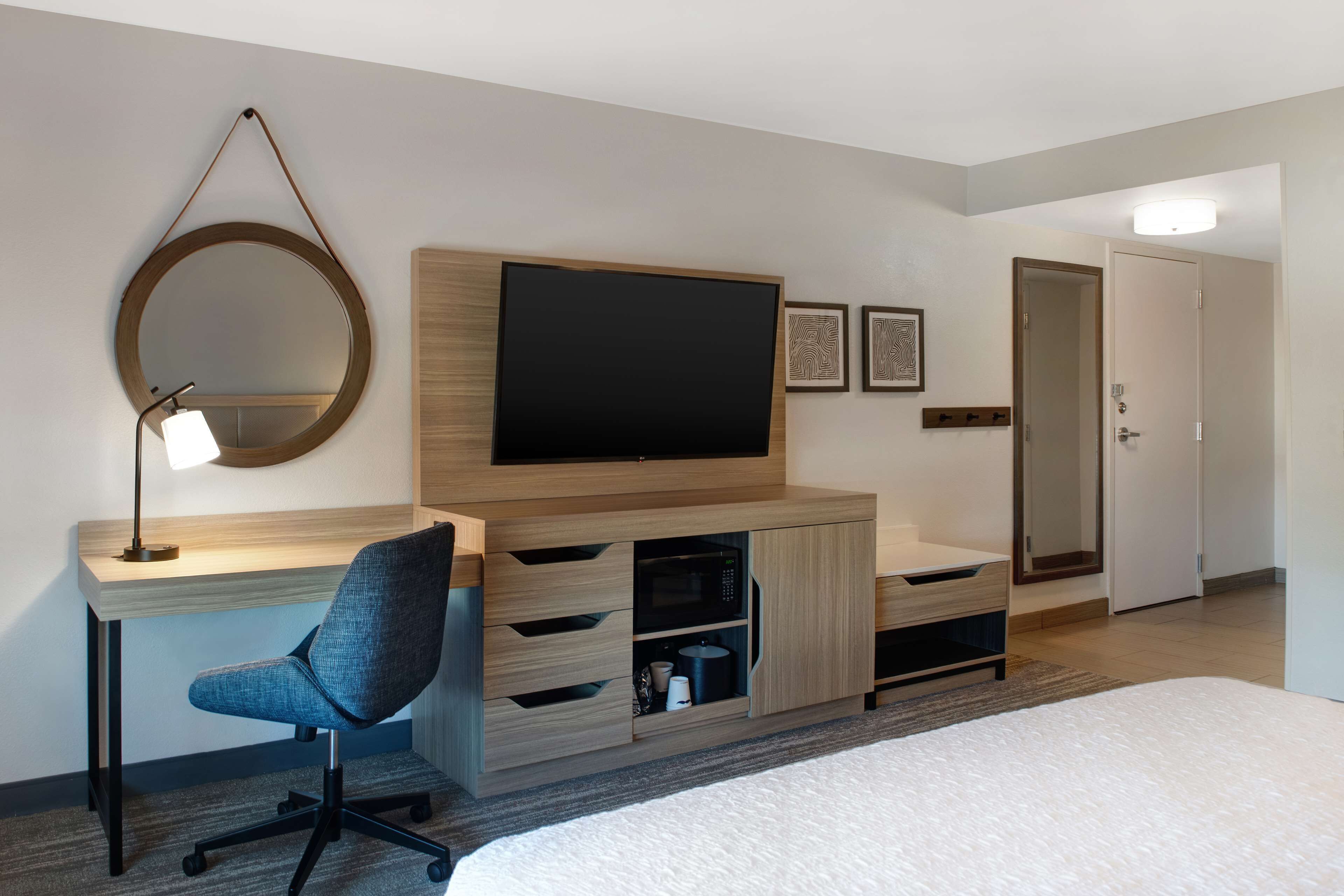 A hotel room with a bed, a desk with a chair, a TV, a mirror, and a door.