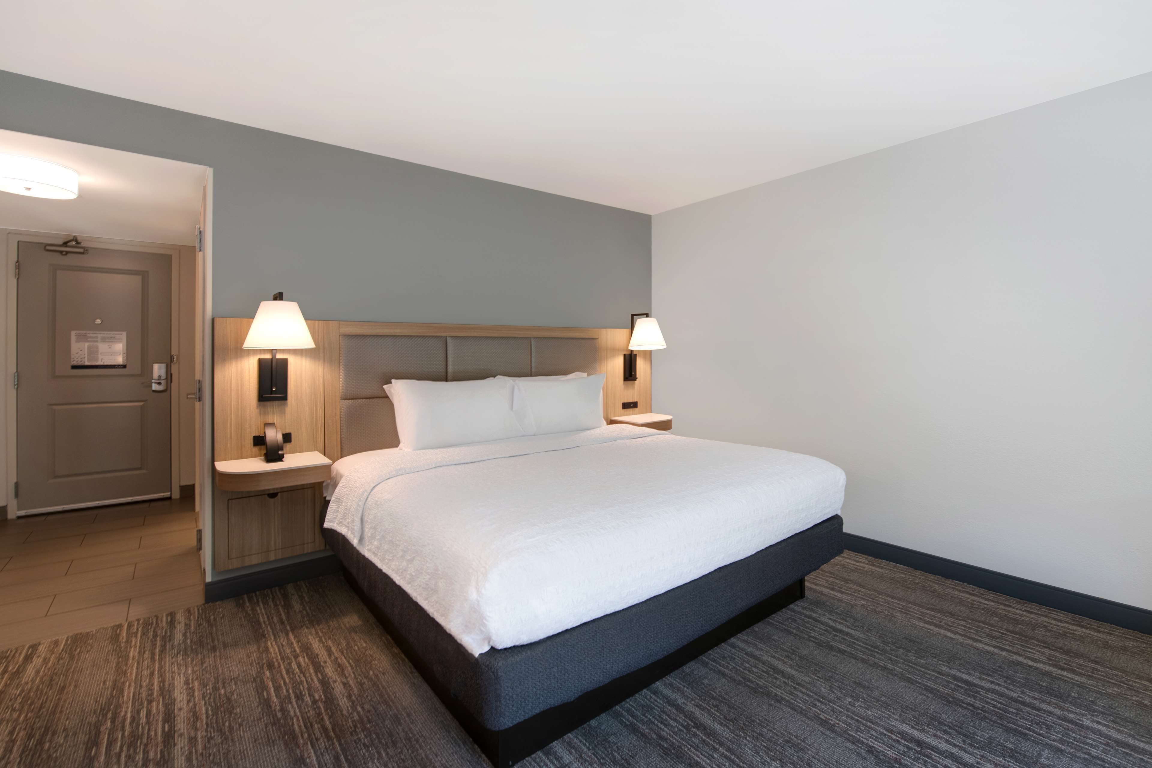 A hotel room with a bed, two bedside lamps, a wooden headboard, a small bedside table, and a carpeted floor.