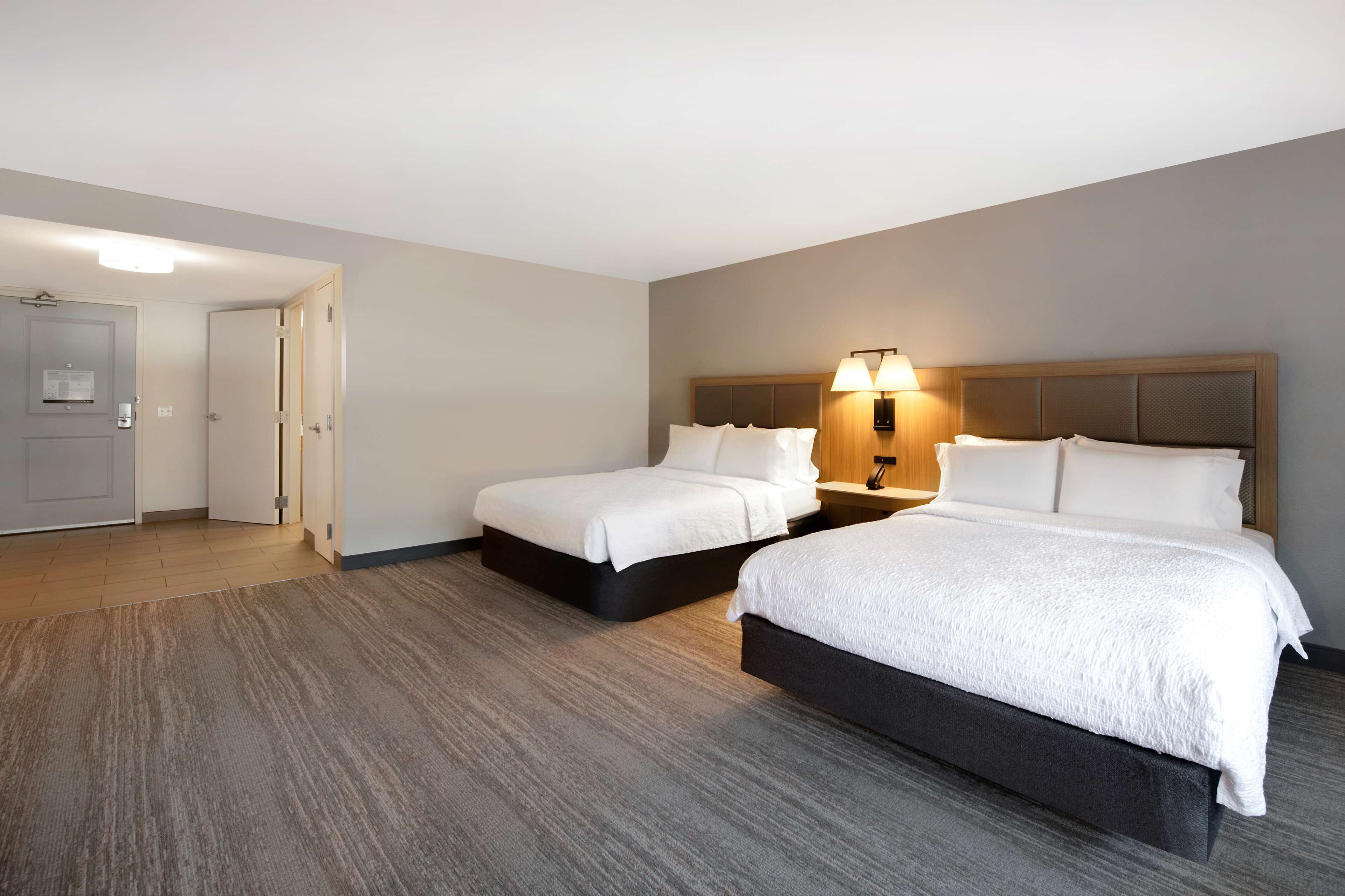 Ein Hotelzimmer mit zwei Betten, Holzboden, einem Nachttisch mit Lampe und einer Tür zu einem weiteren Raum.