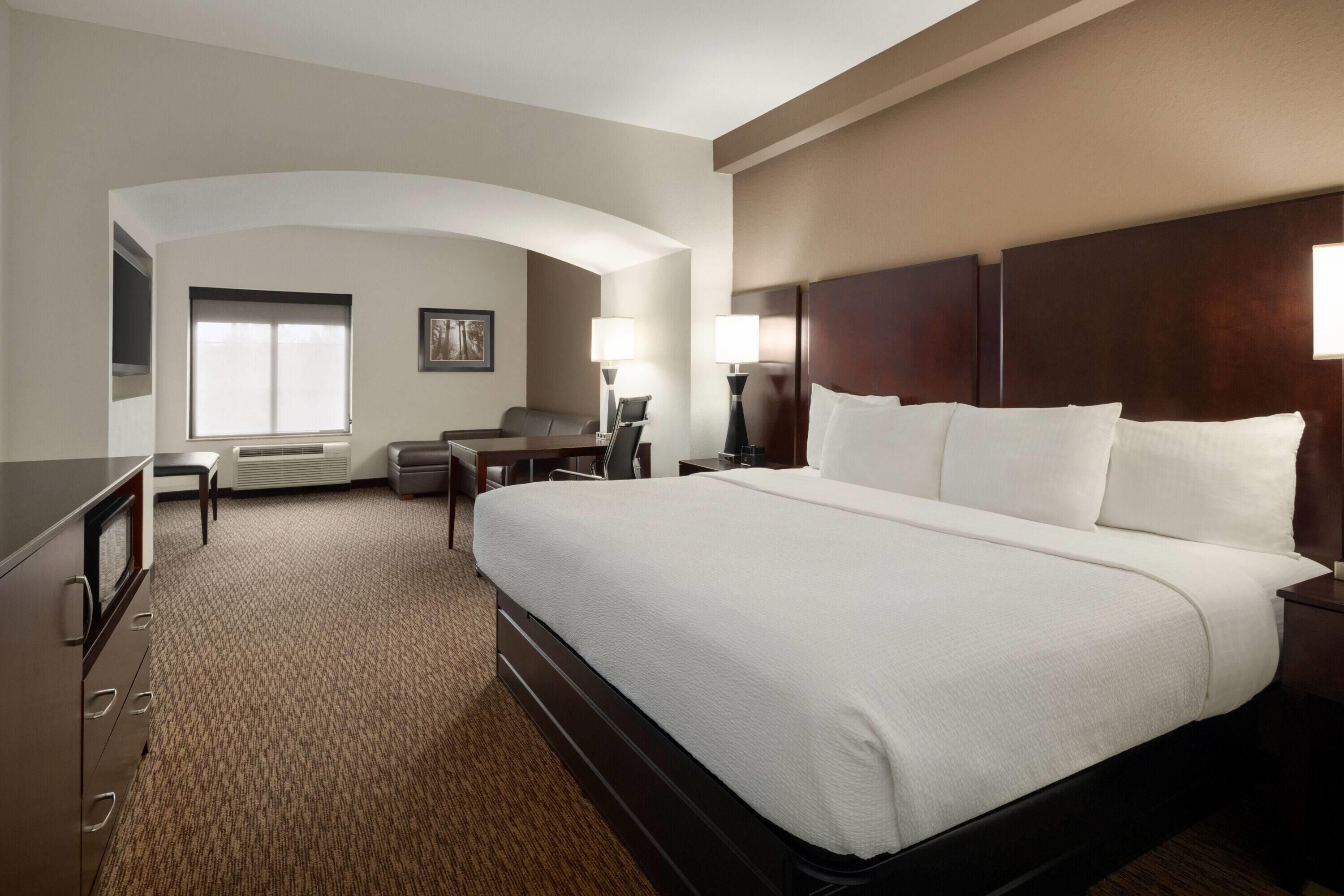 A hotel room with a large bed, a desk, a chair, a lamp, and a window.