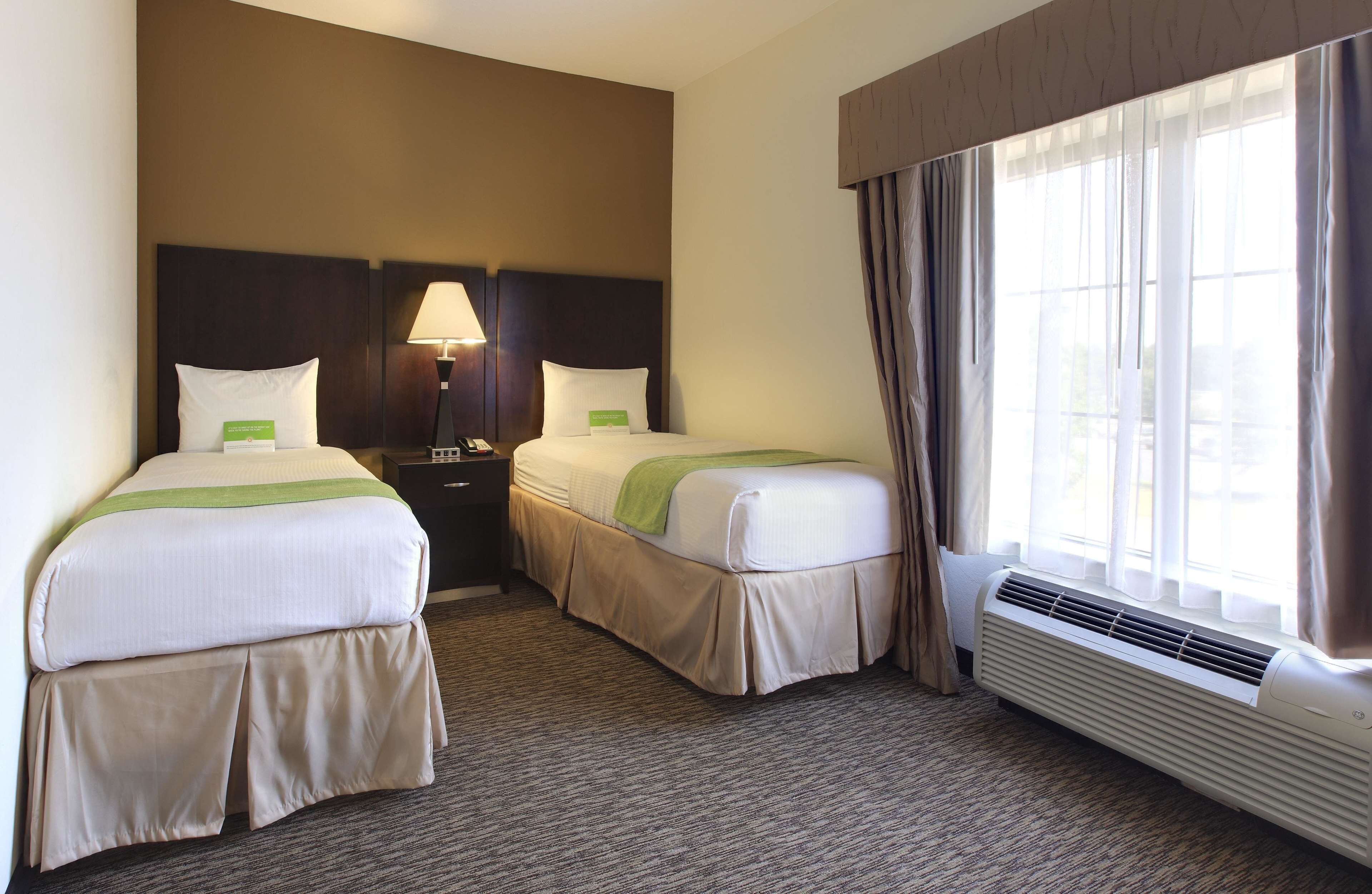 Une chambre d’hôtel avec deux lits, une table de chevet avec une lampe, une fenêtre avec des rideaux et un climatiseur fixé au mur.