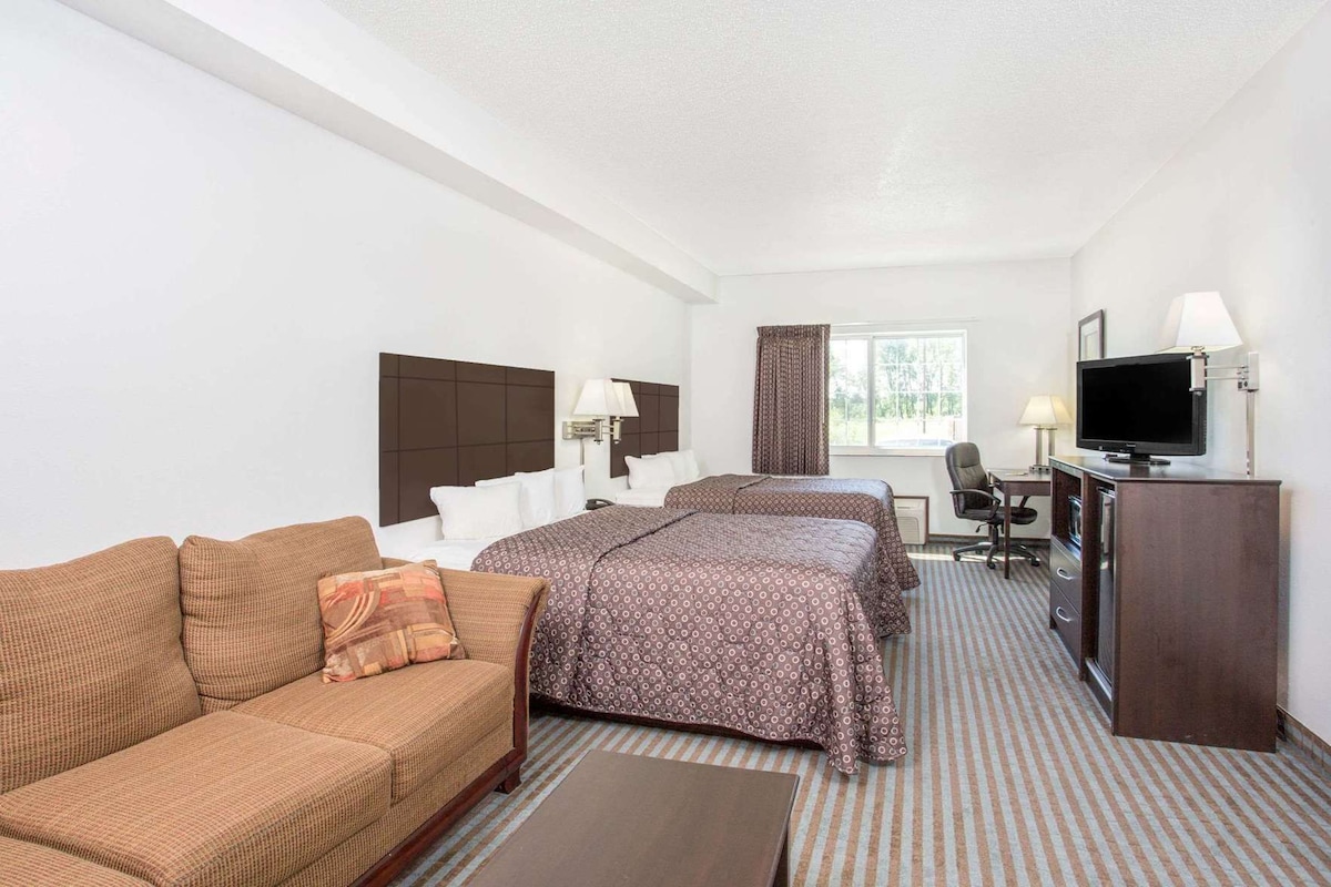 Une chambre d’hôtel avec un canapé, deux lits, un bureau avec une chaise, une télévision et une fenêtre avec des rideaux.