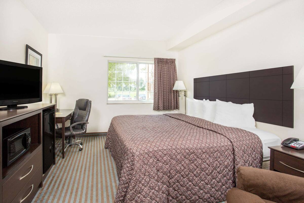 A hotel room with a bed, a desk with a television, a chair, a window with curtains, and a telephone.
