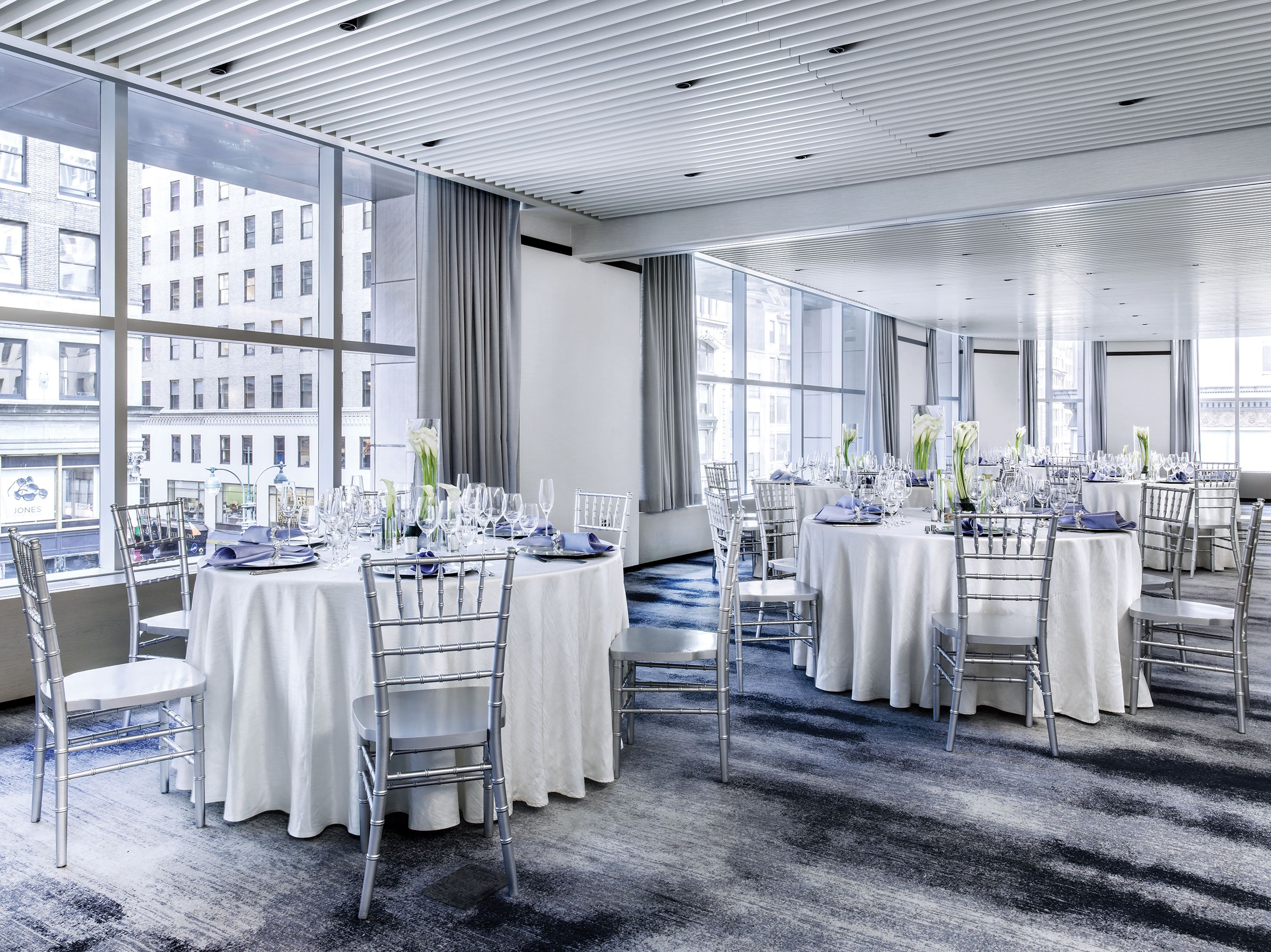 A modern hotel dining area with round tables, white tablecloths, and silver chairs. Large windows offer views of the city.