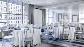 A modern hotel dining area with round tables, white tablecloths, and silver chairs. Large windows offer views of the city.