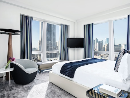 A modern hotel room with a large bed, a flat-screen TV, a city view, and a modern chair.