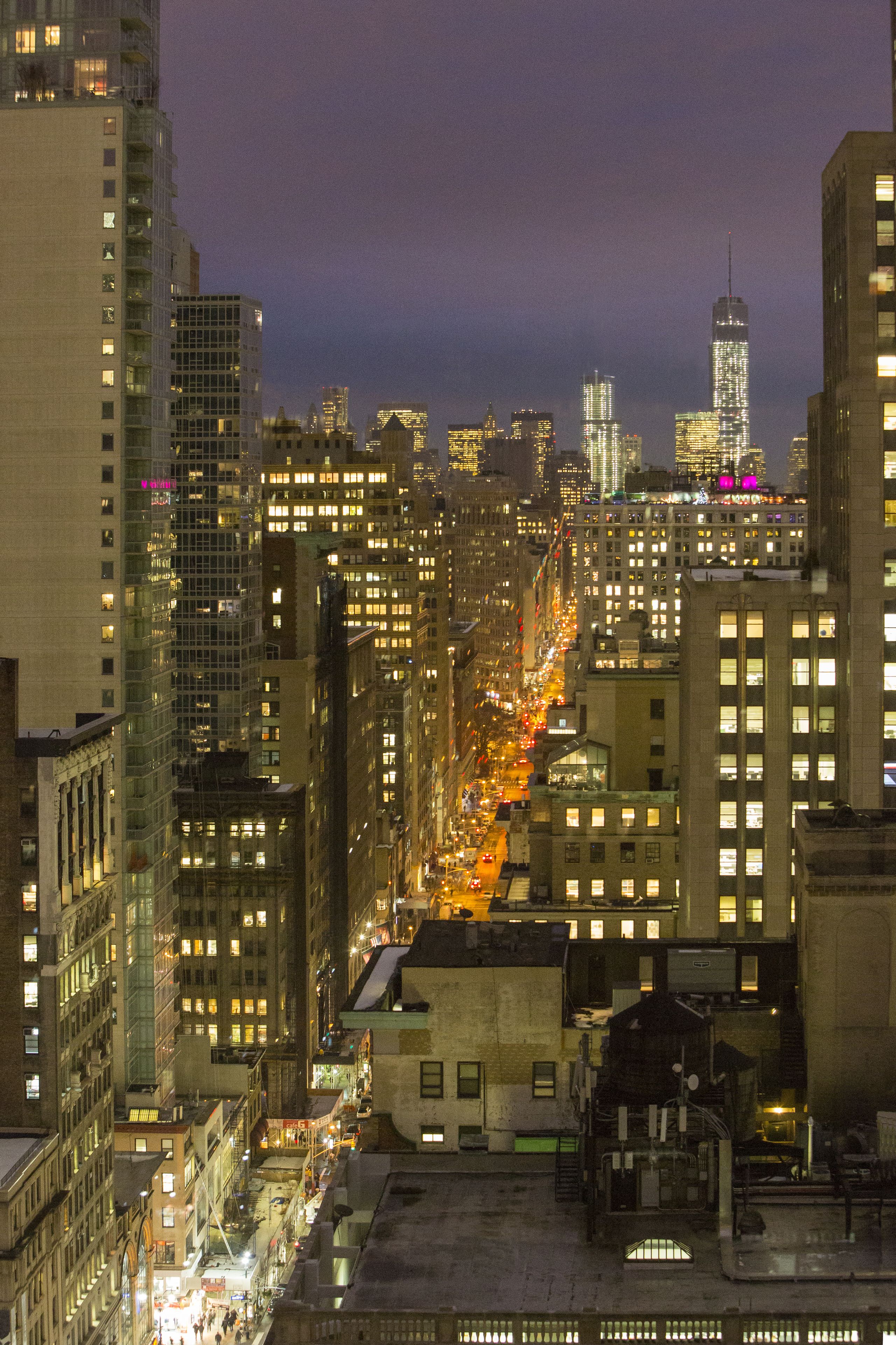 A nighttime cityscape with tall buildings, illuminated windows, and a busy street.