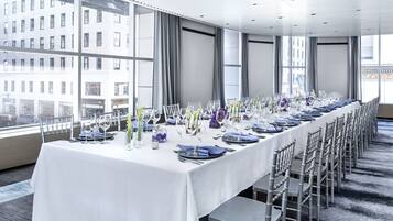A long dining table set for a formal event, with chairs aligned on both sides, large windows offering a city view, and a modern ceiling design.