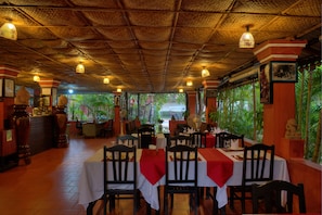 Um restaurante com mesas preparadas para refeições, um telhado de palha e plantas tropicais visíveis pelas janelas.