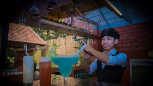 Un espace bar avec un barman qui prépare des boissons. On peut voir différents verres à cocktails sur le comptoir, et un panneau affichant le menu en arrière-plan.