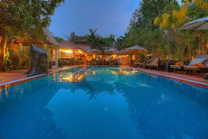 Uma piscina com espreguiçadeiras e guarda-sóis, cercada por árvores e edifícios com ilumina ção quente.