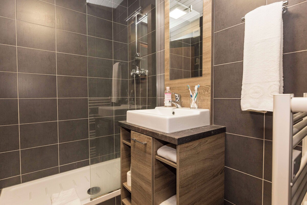 Un baño moderno con baldosas oscuras, ducha envuelta en vidrio, lavabo blanco y tocador de madera.