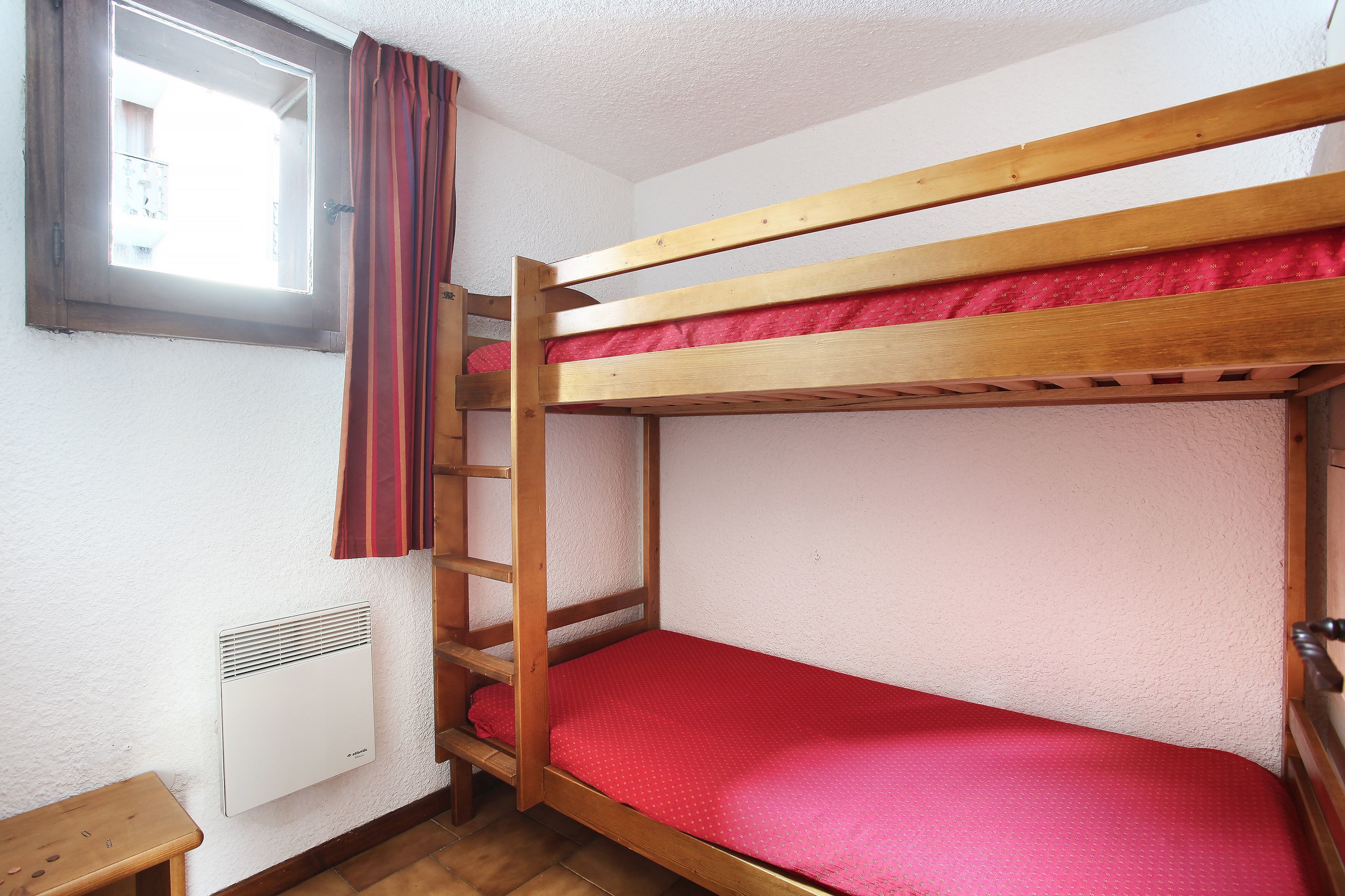 A bunk bed with a red mattress, a window with curtains, and a wall-mounted heater.