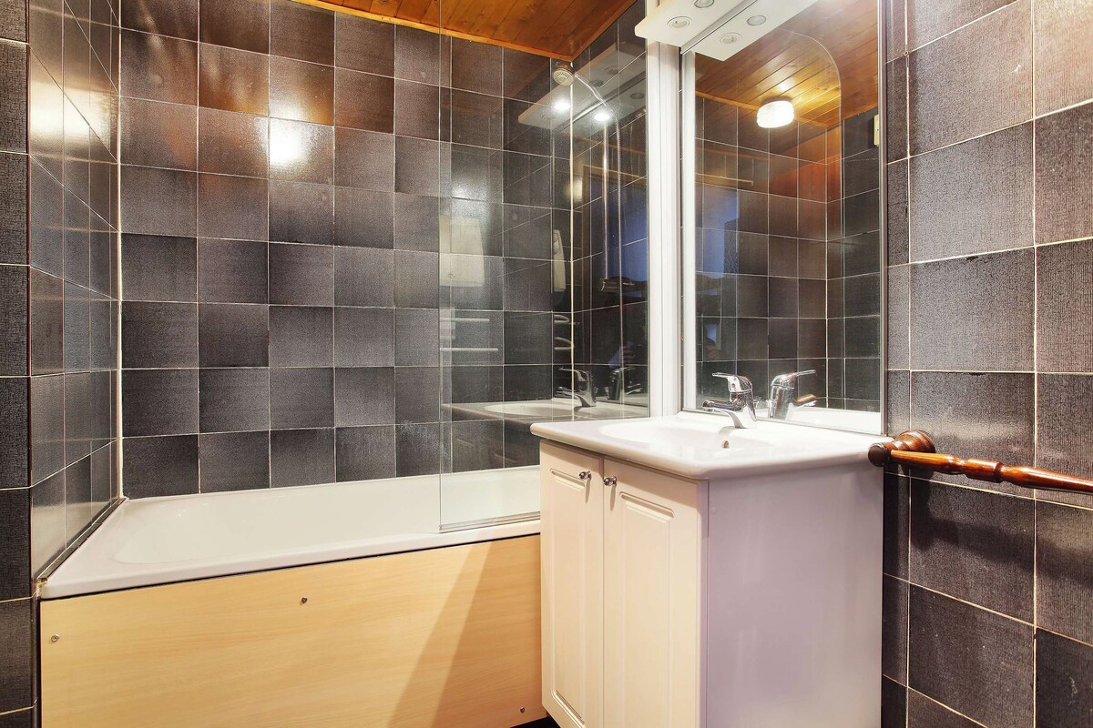 Un baño con paredes de azulejos oscuros, una bañera blanca y un mueble de tocador de madera con espejo.