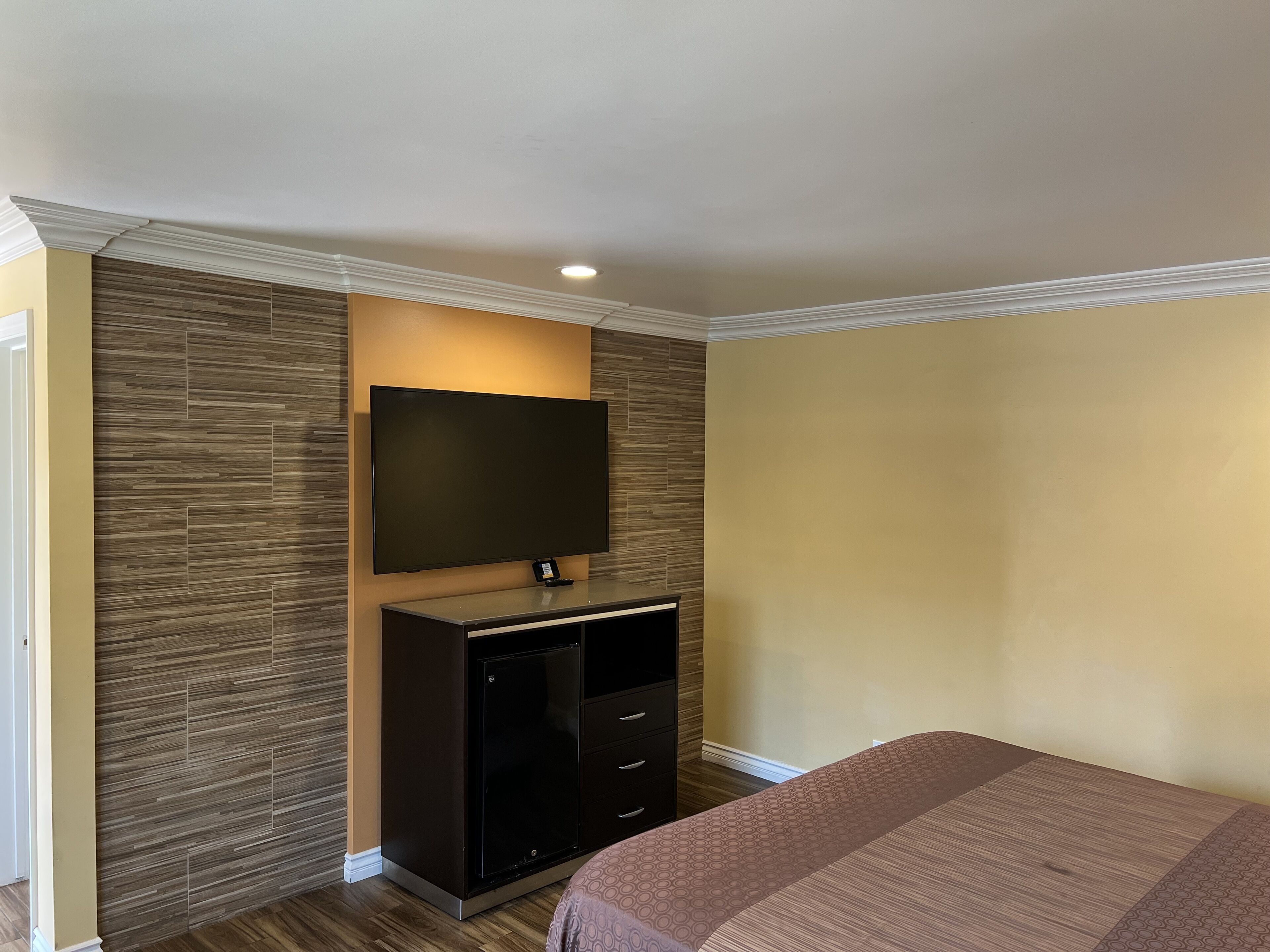 A hotel room with a flat-screen TV mounted on a wall, a dark wooden cabinet below it, and a bed with a patterned bedspread.