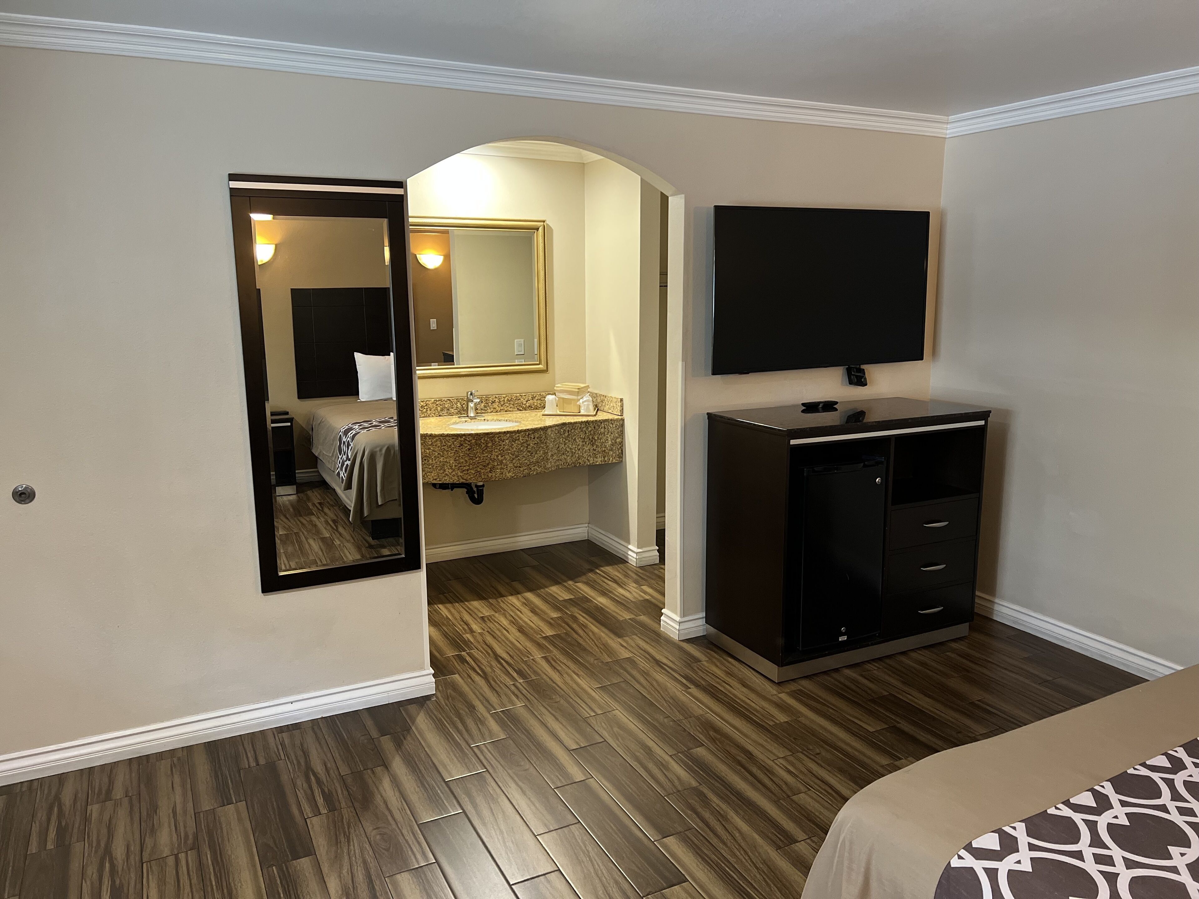 A hotel room with a wooden floor, a bed, a TV mounted on the wall, a vanity with a mirror, and a small cabinet.