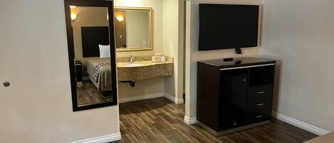 A hotel room with a wooden floor, a bed, a TV mounted on the wall, a vanity with a mirror, and a small cabinet.