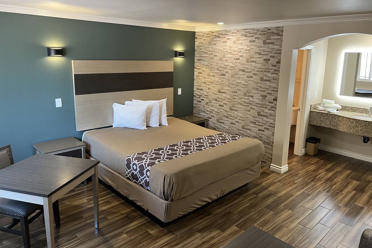A hotel room with a bed, bedside tables, a desk, a bathroom with a sink and mirror, and a wooden floor.