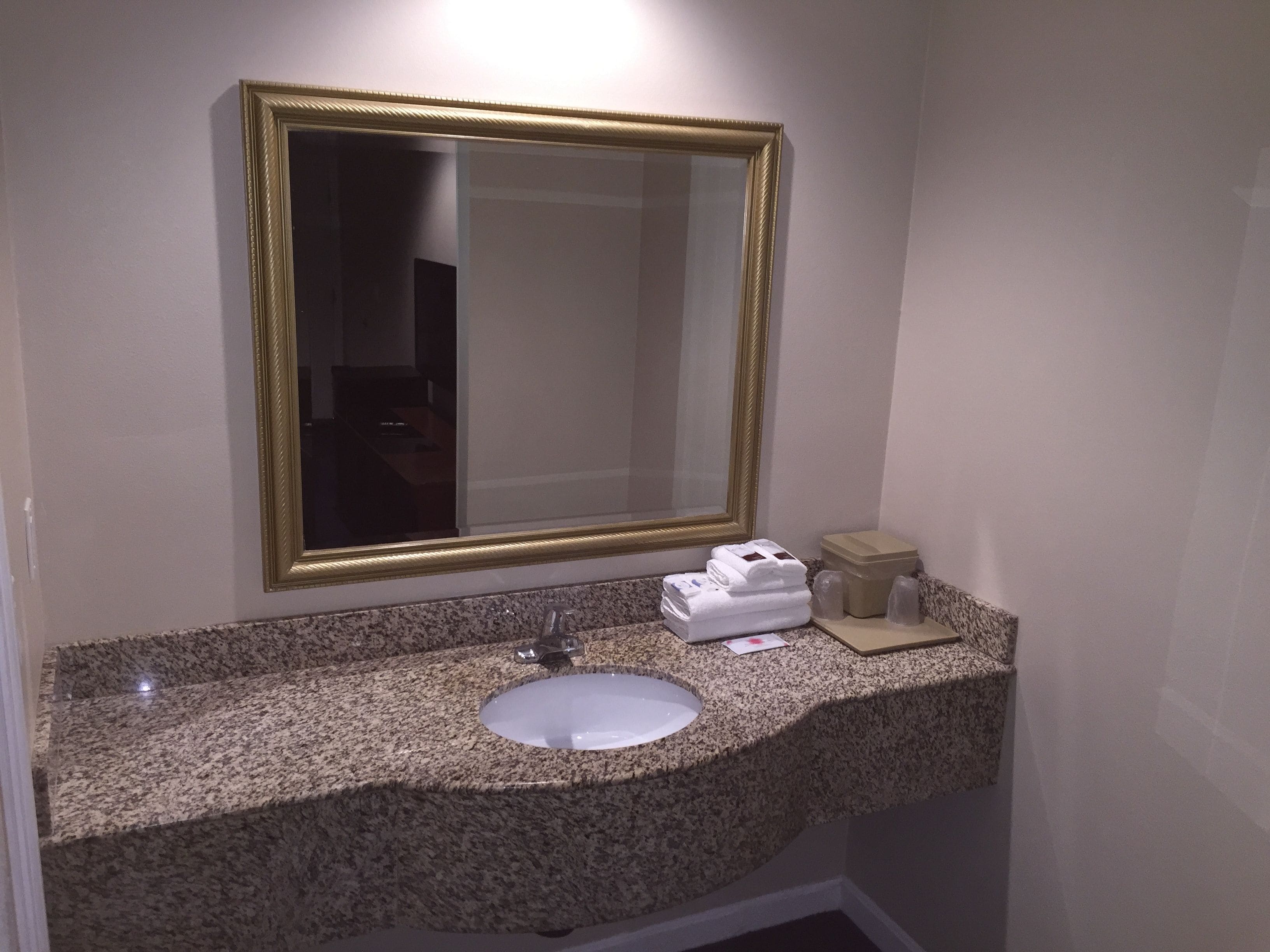 A bathroom with a granite countertop, a white sink, a gold-framed mirror, and a small storage box.